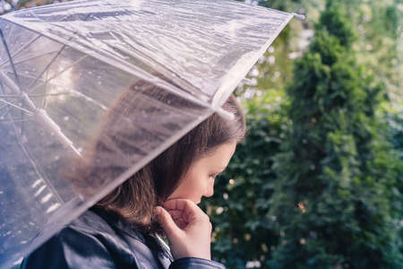 Autumn. Lonely unhappy woman covering her face by hands and cries walking in a park, garden with wet transparent umbrella at rain. Breakup of a couple.の写真素材