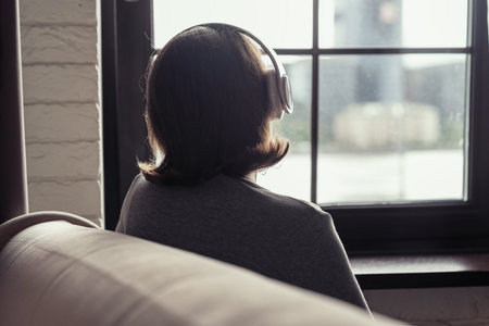 Back view of Ñaucasian young woman in headphones enjoying listening music sitting near window of...