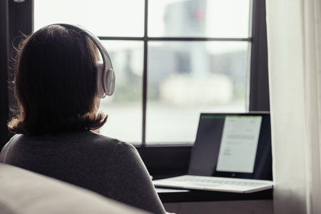 Back view of lonely woman listenning music in headphones and working on laptop sitting near window...