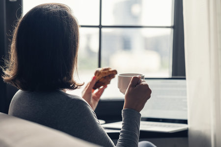 Back view of lonely woman enjoying with cup of coffee and croissant working on laptop sitting near...