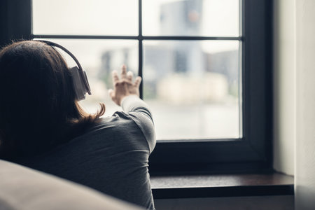Back view of lonely brunette woman in headphones listen music and touching window. Conceptの写真素材
