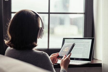 Woman holding american passport using laptop for making order and booking sitting near window in the...