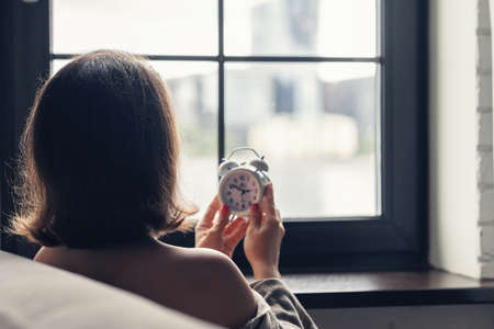 Back view of lonely caucasian young woman wrapped in a plaid with with alarm clock sitting near window at cold winter day.の写真素材