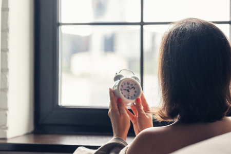 Back view of lonely caucasian young woman wrapped in a plaid with with alarm clock sitting near...