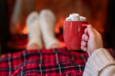 Woman with cup of hot cocoa and marshmallow warming legs in winter white socks near fireplace flame, covered christmas plaidの写真素材