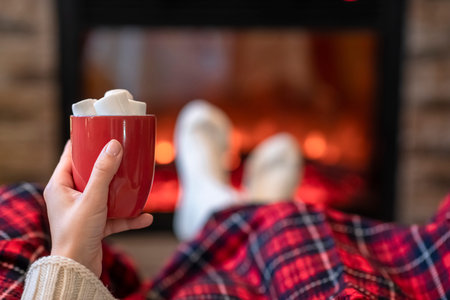 Woman with cup of hot cocoa and marshmallow warming legs in winter white socks near fireplace flame,...