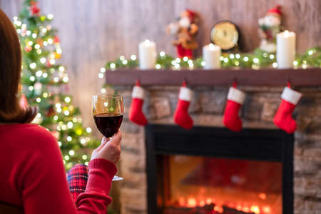 Woman hand with glass of wine covered plaid sitting and relaxation on armchair near christmas fareplace and fir-tree.の写真素材