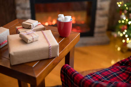 Armchair covered plaid, fareplace and christmas tree. Cup of cocoa with marshmallows packing gift boxes for family on table.の写真素材