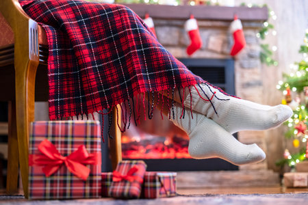 Woman legs in a winter socks covered plaid sitting and relaxation on armchair near fireplace and...
