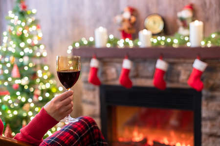 Woman hand with glass of wine covered plaid sitting and relaxation on armchair near christmas fareplace and fir-tree.の写真素材