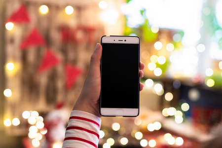 Woman holding mobile phone and showing screen with christmas lights background. Concept of online service in holidays.の写真素材