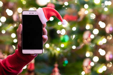Woman holding mobile phone with santa claus red hat showing screen with christmas tree lights on the background. Concept of online service in holidays.の写真素材