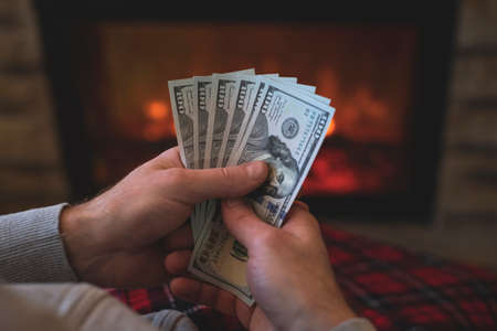 Man counting american dollars sitting near christmas tree and fireplace. Spending money on gift at christmas time. concept. Back view.の写真素材