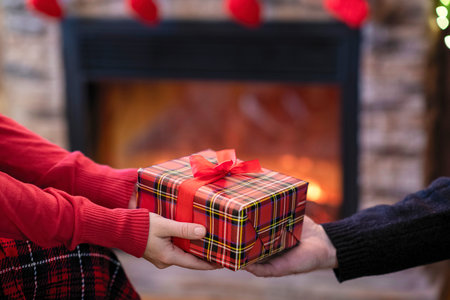 Man giving Christmas and New Year Gift box to woman in front of the fireplace with flame.