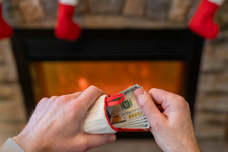 Man putting american dollars in red christmas sock sitting near christmas tree and fireplace....