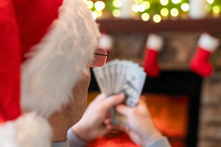 Man in a Santa claus hat counting american dollars sitting near christmas tree and fireplace....