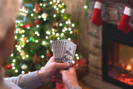 Man counting american dollars sitting near christmas tree and fireplace. Spending money on gift at christmas time. concept. Back view.の写真素材