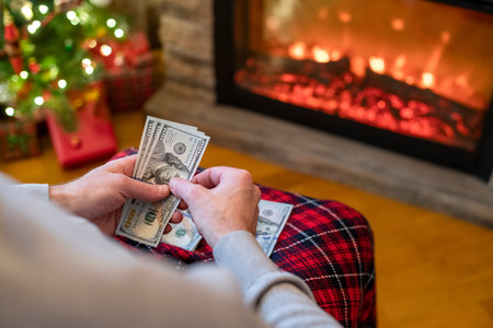 Man counting american dollars planning trip sitting near christmas tree and fireplace. Spending...