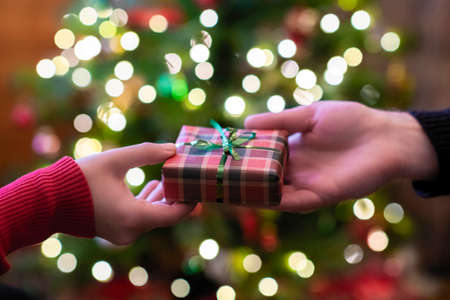 Man giving Christmas and New Year Gift box to woman opposite christmas tree with glowing light bulbs. Xmas concept. Close up. Selective focusの写真素材