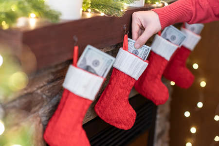Woman putting american dollars in Ñhristmas red socks for gifts on the fireplace in bokeh lights.の写真素材