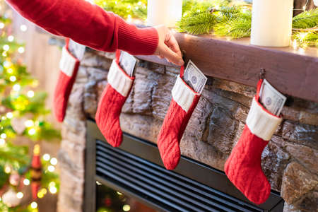 Woman putting american dollars in Ñhristmas red socks for gifts on the fireplace in bokeh lights.の写真素材