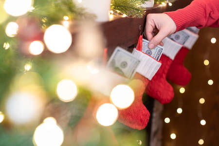 Woman putting american dollars in Christmas red socks for gifts on the fireplace in bokeh lights.の写真素材