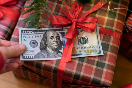 Woman putting american dollars under red bow of huge gift box lying under christmas tree. Concept