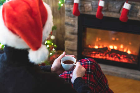 One divorce man with cup coffee, tea in santa claus hat sitting and warming at winter evening near fireplace flame, covered christmas plaidの写真素材