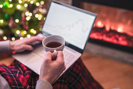 Man freelancer working on laptop with graphs and charts on screen sitting near christmas tree and fireplace with cup of tea.の写真素材