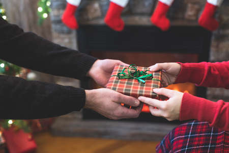 Man giving Christmas and New Year Gift box to woman in front of christmas tree, fireplace with flame. Xmas concept.の写真素材