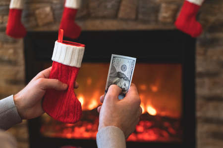 Man putting american dollars in red christmas sock sitting near christmas tree and fireplace. concept.の写真素材
