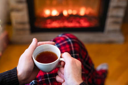 Man with cup coffee, tea in santa claus hat sitting and warming at winter evening near fireplace flame, covered christmas plaidの写真素材