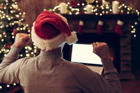 Christmas. Man in santa claus hat with laptop sitting near christmas tree and fireplace. Concept of...