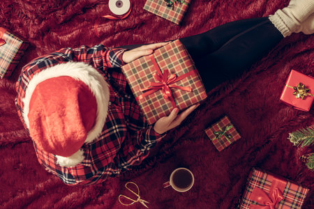 Top view of woman with christmas gift box on knees. Female wrapping gifts for family sitting on fluffy plaid near christmas tree. Flat lay.の写真素材