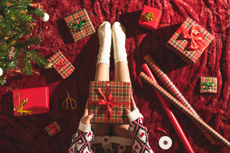 Top view of woman with christmas gift box on knees. Female wrapping gifts for family sitting on fluffy plaid near christmas tree. Flat lay.の写真素材