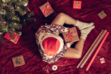 Top view of woman in red santa claus hat wrapping christmas gifts boxes for family sitting on fluffy plaid near christmas tree. Flat lay.の写真素材