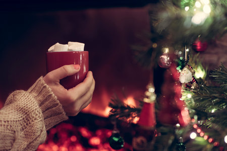 Woman hand with cup of hot cocoa and marshmallow sitting and warming at winter evening near fireplace flame. View through the branches of a Christmas treeの写真素材