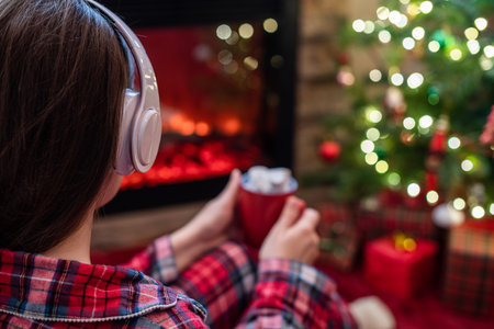 Woman in pajama in headphones with cup of hot cocoa and marshmallow sitting and warming at winter evening near fireplace flame and christmas tree.の写真素材