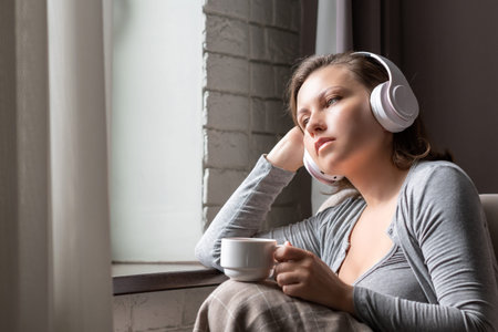 Lonely sad beautiful woman with cup of coffee listenning music in headphones sitting on sofa near...