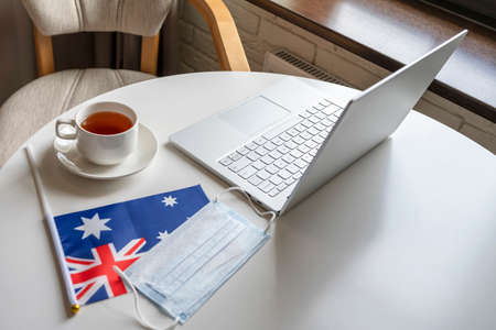 Woman tourist having breakfast with cup of coffee working on laptop. Flag of Australia, medical protective face mask on table of cafe. Protection from bacteria and viruse in a public place. Concept.の写真素材