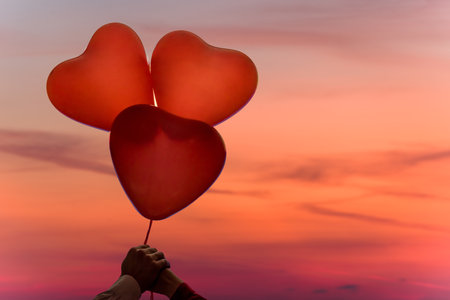Valentines day. Silhouette of man and woman hands holding three reds balloons in form of heart on pastel sunset pink sky background. Beach. Conceptの写真素材