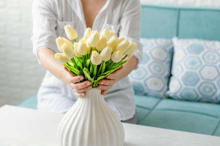 Woman putting white tulips flowers in vase sitting at the living room coffee table. Composing bouquet. Lifestyleの写真素材