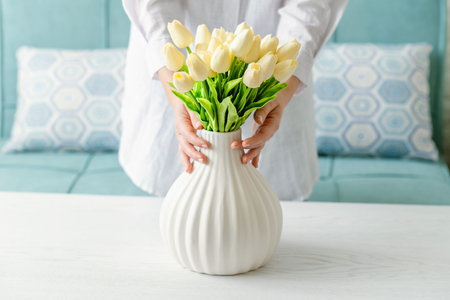 Woman putting white tulips flowers in vase sitting at the living room coffee table. Composing...