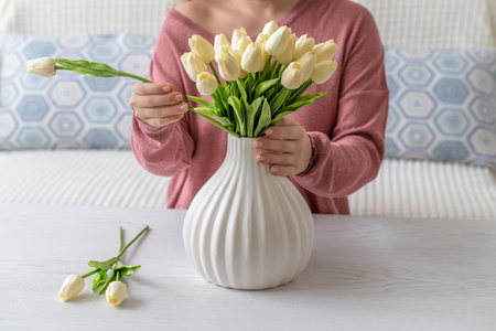 Woman putting white tulips flowers in vase sitting at the living room coffee table. Composing...