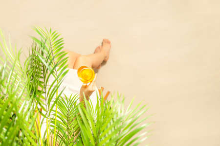 Alone woman sitting under palm tree branches with glass of water with piece orange. Female relaxation on the sand of the beach at summer vacation. Top viewの写真素材