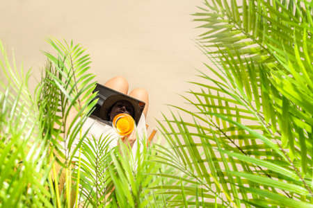 Woman blogger with tablet holding glass of water with piece orange sitting under palm tree branches on the sand of beach. Summer vacation. Top viewの写真素材