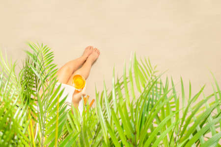 Alone woman sitting under palm tree branches with glass of water with piece orange. Female relaxation on the sand of the beach at summer vacation. Top viewの写真素材