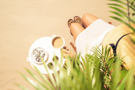 Woman on the beach drinking hot coffee with chocolate cookies sitting under palm trees at table and enjoying during exotic summer vacation. Outdoors cafe. Top viewの写真素材