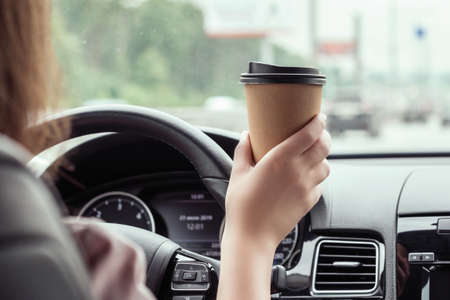 woman is driving on the highway and holding paper cup of coffee at the morning. View from the back seat of the carの写真素材