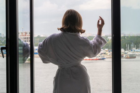 Back view of beautiful young woman in bath terry robe with cup of coffee standing and enjoying near...